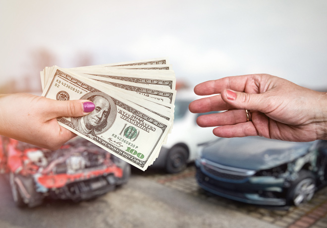 A woman's hand paying several hundred dollars in cash to another hand with cars in the background.